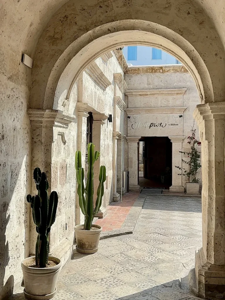 A sunlit stone archway and corridor in a hotel courtyard, featuring potted cacti and leading to another arched opening and a building with a "Hampton by Hilton" sign in Arequipa.