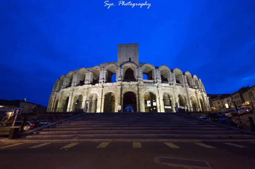 The illuminated Roman Amphitheatre in Arles at dusk, showcasing its grand arched facade and imposing structure against a deep blue sky.