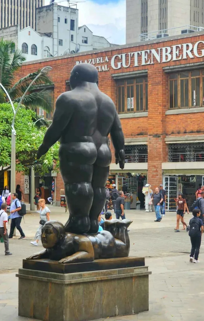 A large bronze "Fat Man" sculpture by Fernando Botero standing on a reclining figure in Botero Plaza, Medellín, with a red brick building in the background.