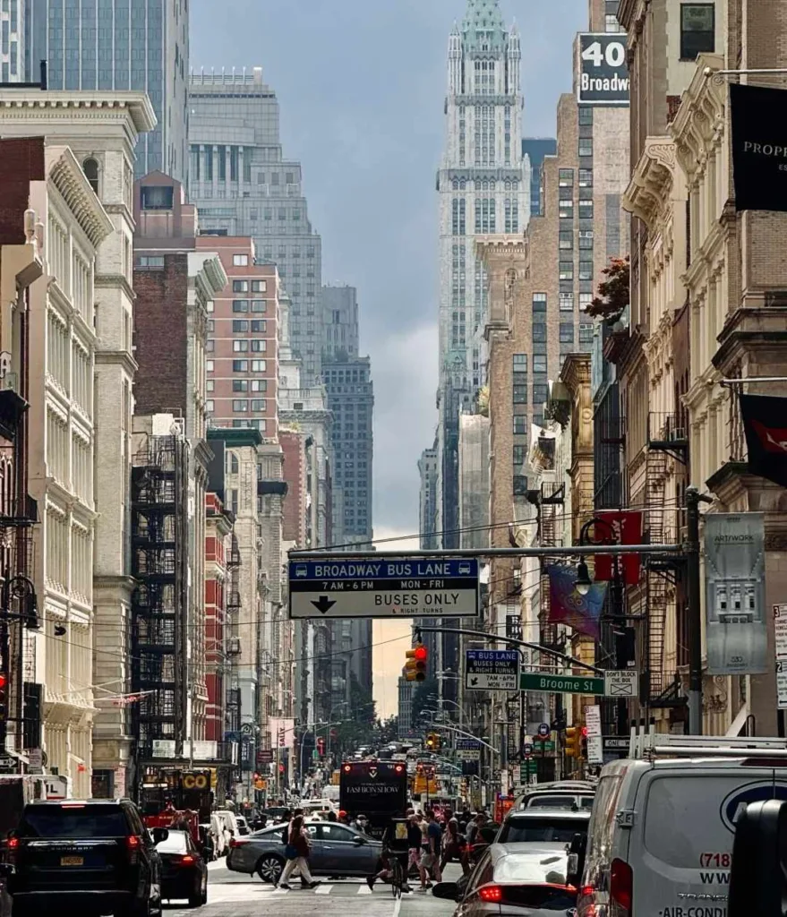 A view down a busy Broadway street in New York City, flanked by tall, varied buildings and featuring heavy traffic, pedestrians, and overhead street signs.