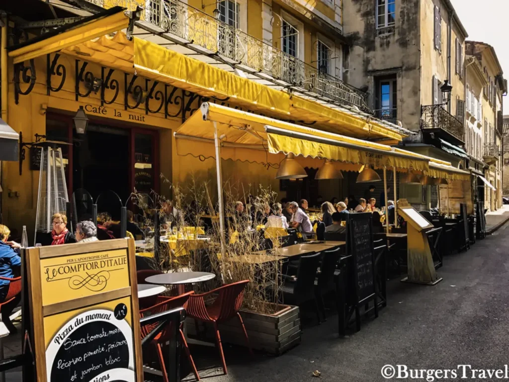 The real-life Cafe Van Gogh in Arles, France, with its distinctive yellow facade and awnings, outdoor tables and chairs filled with patrons, on a sunny day.