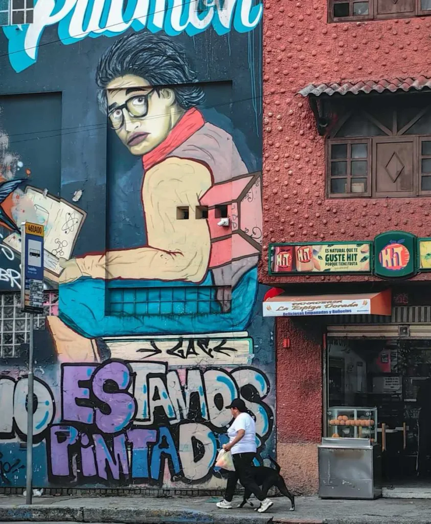 A vibrant street art mural in Bogotá depicting a person with glasses and a book, covering the side of a building next to a shop.