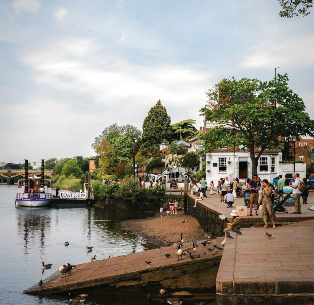 Charming English riverside town with boat trips sign, locals gathered by water with ducks and peaceful atmosphere
