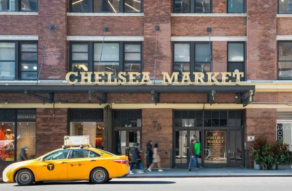 The brick facade entrance of Chelsea Market in New York City, with its distinctive sign, a yellow taxi, and pedestrians on the sidewalk.