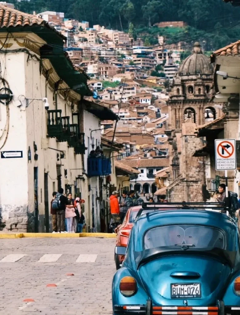 A vibrant street scene in Cusco, Peru, with traditional colonial-style buildings and people on a sloping cobblestone road, leading up to a distant hillside covered with houses. A blue vintage car is parked in the foreground.