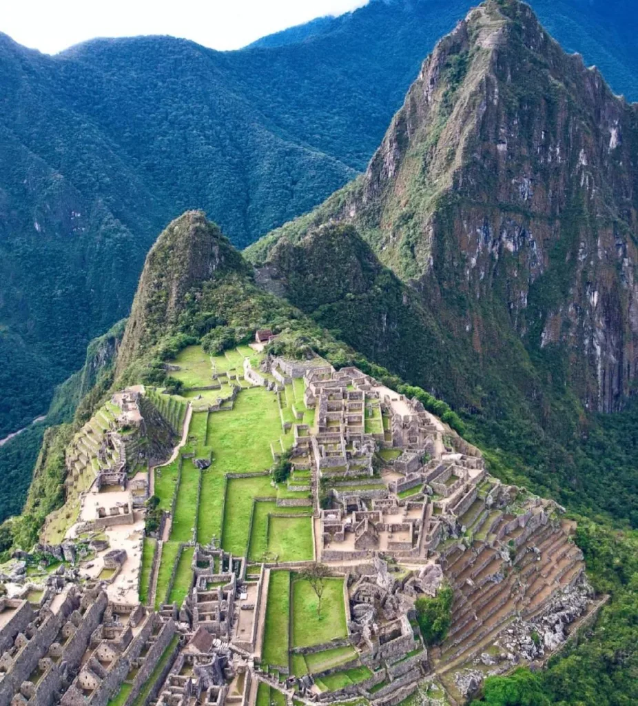 An aerial view of the ancient Inca city of Machu Picchu, with stone structures and green terraces nestled between towering, lush green mountains under a cloudy sky.