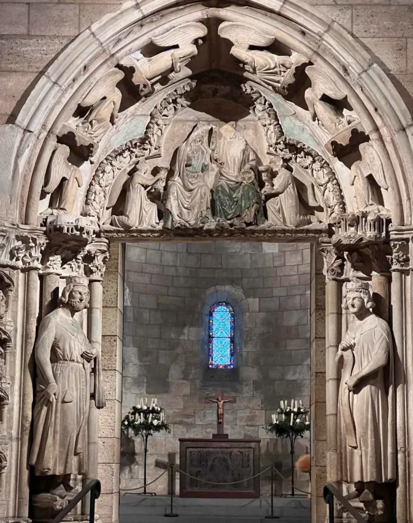 A detailed medieval stone archway inside The Cloisters Museum, with religious sculptures and a small, colorful stained-glass window at the end of a corridor.