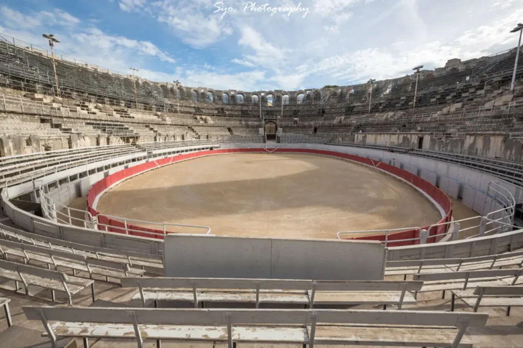The vast, empty interior of the ancient Roman Amphitheatre in Arles, with tiered seating surrounding a large central arena, under a partly cloudy sky.