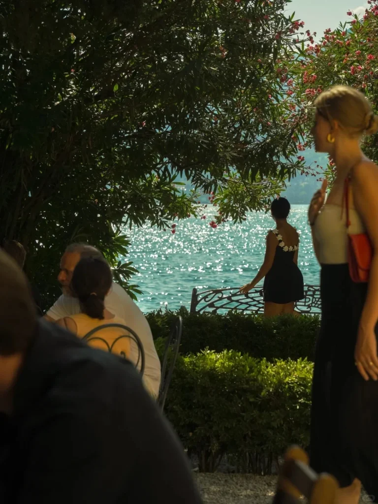 People sitting at an outdoor cafe, with a woman in a dark dress standing by a decorative railing overlooking the shimmering, sunlit turquoise water of a lake. Lush green trees and flowering bushes frame the view.