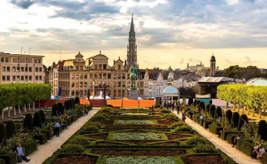 Brussels cityscape with Gothic church spire, formal geometric gardens, and historic guild houses under dramatic cloudy sky
