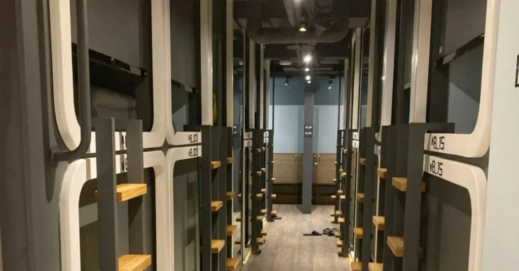 A long, narrow hallway lined with modern, individual capsule beds with wooden shelves and white frames, indicating a capsule hotel or hostel dormitory.