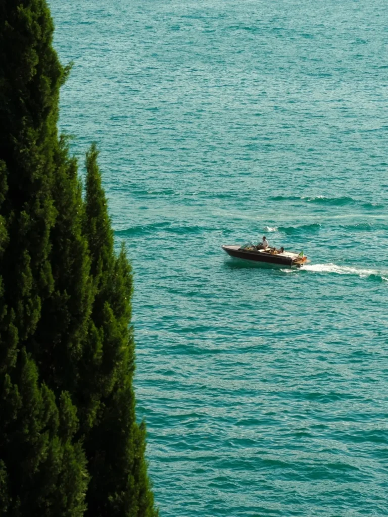 A classic wooden motorboat with two people on board speeds across the turquoise-blue, rippling water of a lake, with a tall, dark green cypress tree partially visible on the left.