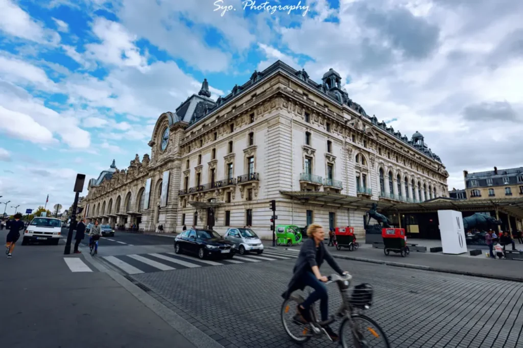 The grand exterior of the Musée d'Orsay, a former railway station, with a cyclist and other people on the street in Paris under a partly cloudy sky.