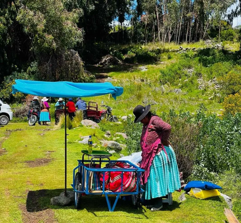 A person wearing a hat and traditional skirt standing next to a small blue vending cart with a blue umbrella for shade, in a grassy, rural area with trees and other people in the background.