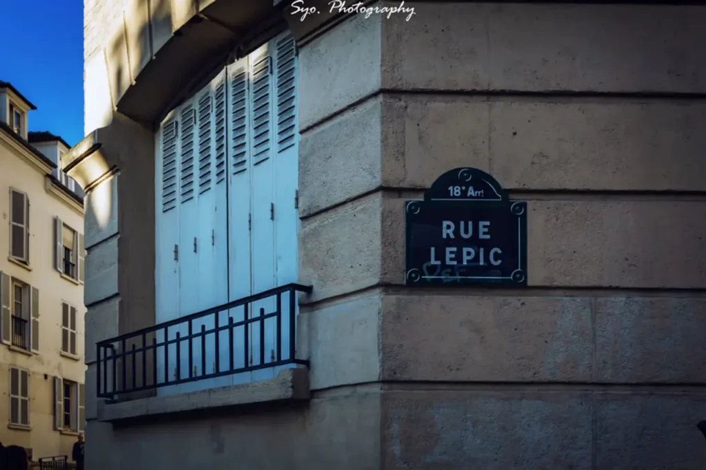 A close-up of a blue street sign with white text that reads "RUE LEPIC" and "18e Arr" (18th Arrondissement), mounted on a stone building corner with a balcony.