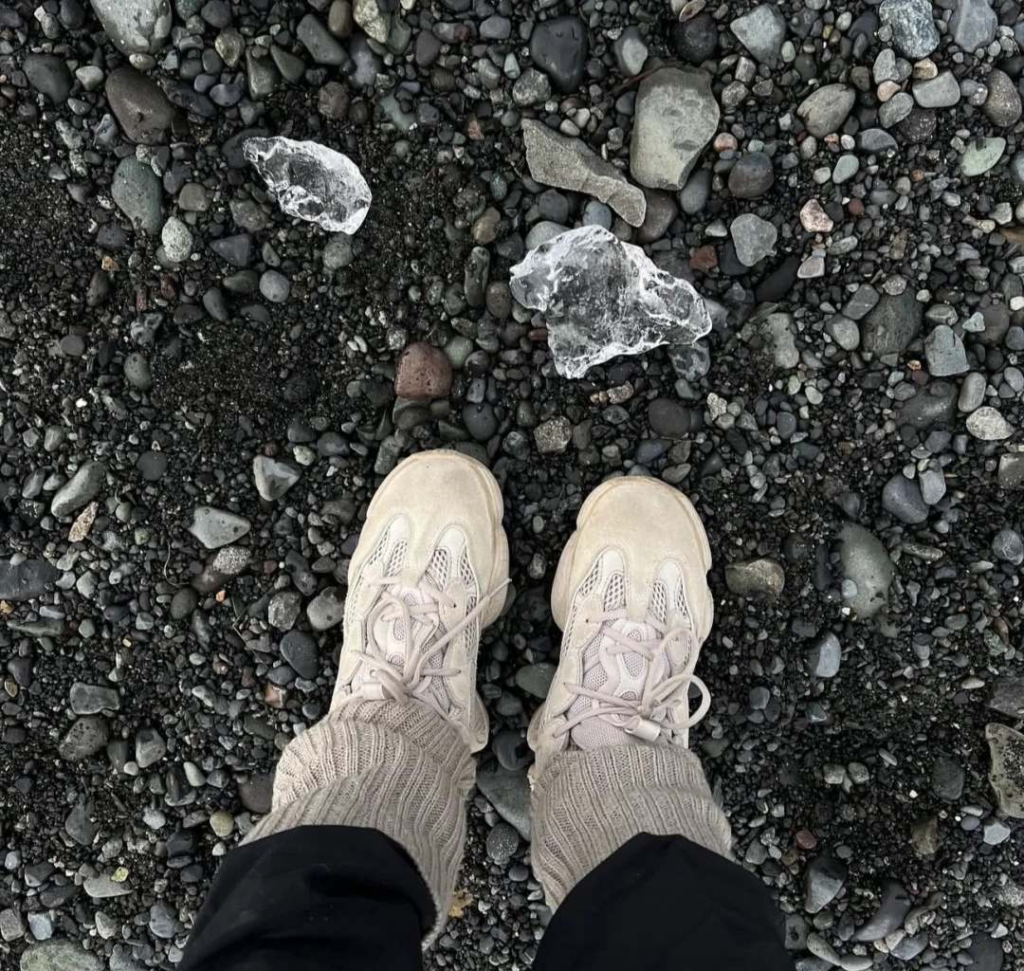 Solo traveler's feet in sneakers standing on black volcanic beach with clear ice chunks and pebbles