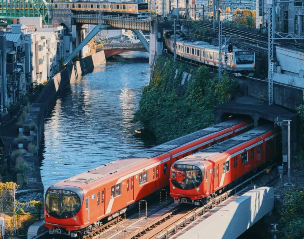 Two red Tokyo subway trains on tracks next to a river, with multiple bridges and other train lines visible above and in the background, under a blue sky.