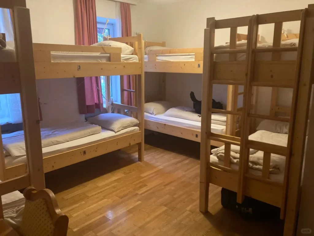 A bright hostel dorm room with multiple wooden bunk beds, white bedding, and a light wooden floor, with a window and pink curtains visible in the background.