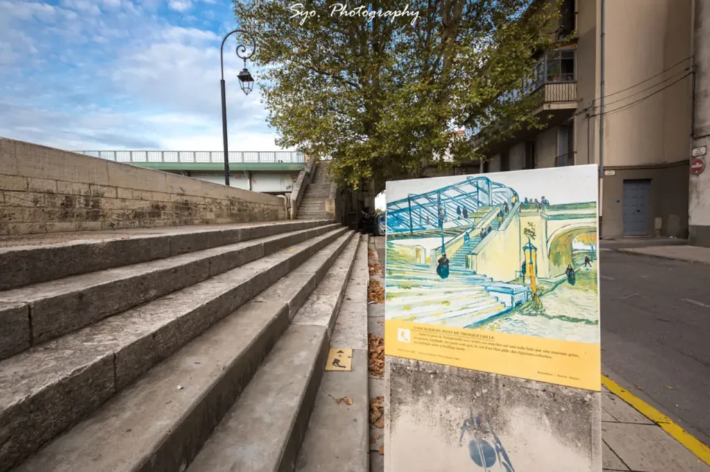 The stone steps leading up to the Trinquetaille Bridge in Arles, with a large reproduction of Van Gogh's "The Trinquetaille Bridge" painting displayed prominently in the foreground.
