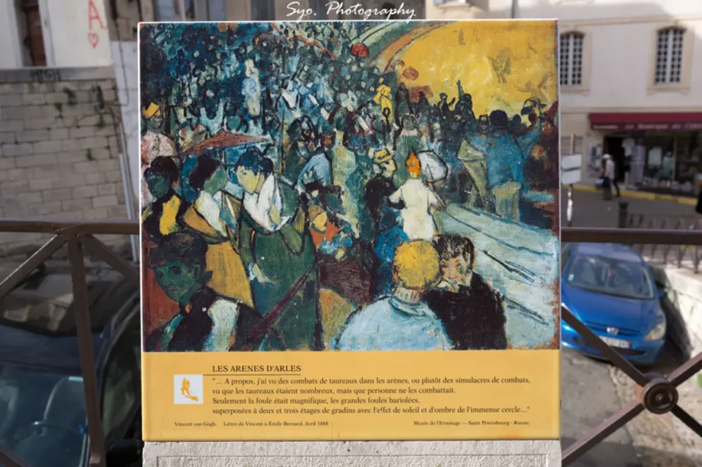 A framed reproduction of Vincent van Gogh's painting "Les Arènes" (The Arena at Arles), showing a vibrant crowd in an amphitheater, displayed outdoors with a street scene in the background.