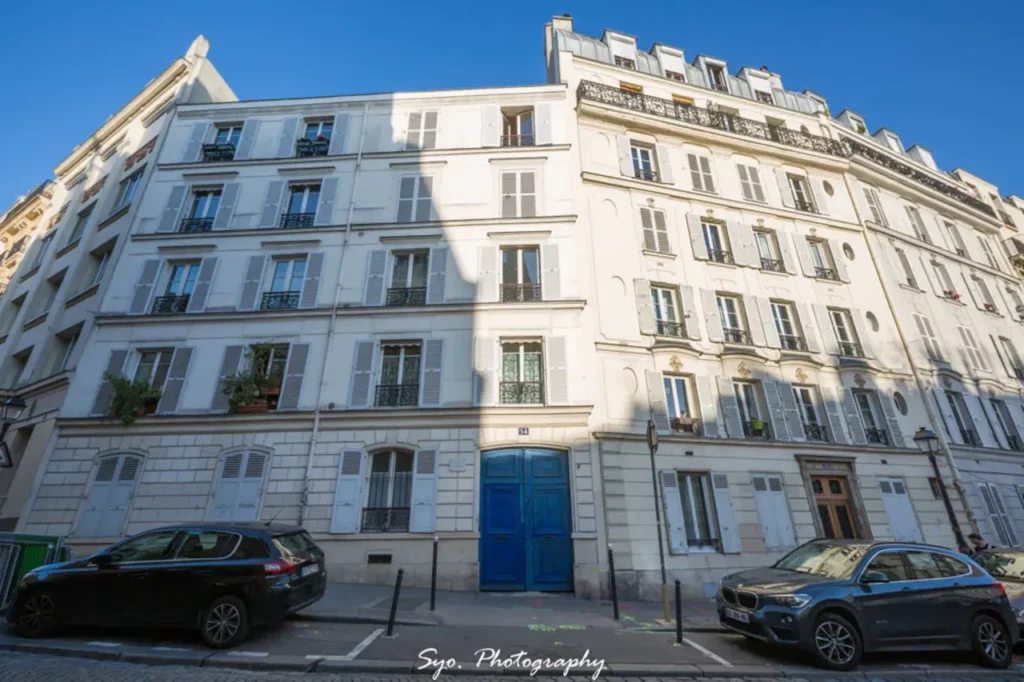 A large, multi-story Parisian apartment building with light-colored facades, numerous windows with shutters, and a distinctive blue entrance door on Rue Lepic. Two cars are parked in front.