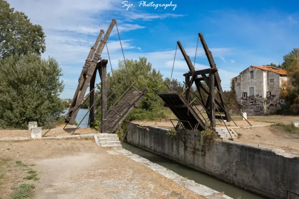 The wooden drawbridge known as Langlois Bridge (Pont de Langlois) in Arles, with its two arms raised, surrounded by green trees and a disused building under a blue sky with white clouds.
