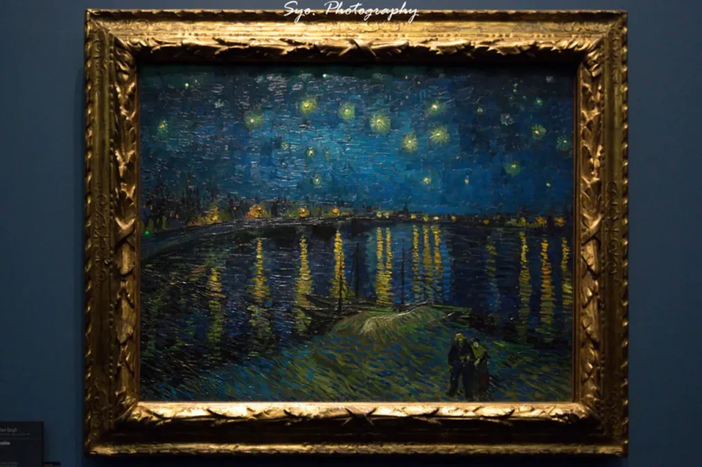 A framed oil painting by Vincent van Gogh, depicting a starry night sky over the Rhône River with city lights reflecting on the water, and two figures in the foreground.