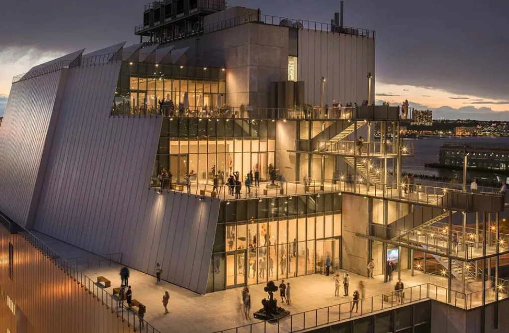 he modern, multi-level exterior of the Whitney Museum of American Art in New York City at dusk, with lights illuminating its terraces and large windows showcasing visitors inside.