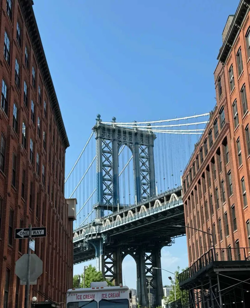 Manhattan Bridge framed by historic red brick buildings in DUMBO Brooklyn with blue sky and ice cream truck