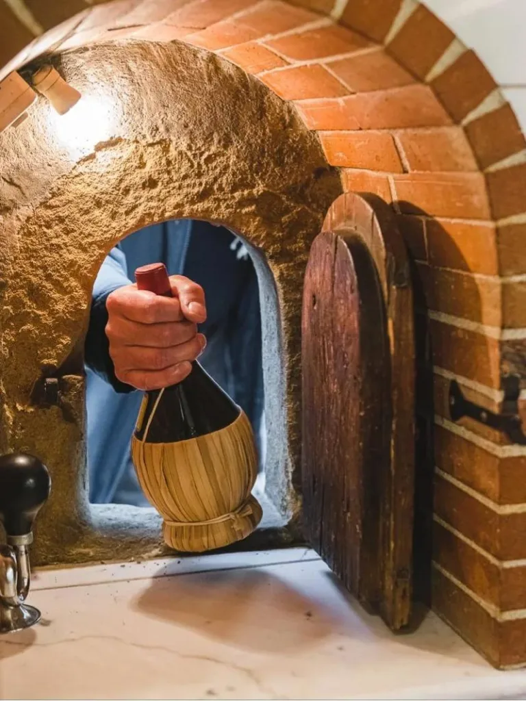 Hand holding traditional Tuscan fiasco wine bottle through arched stone wine window opening