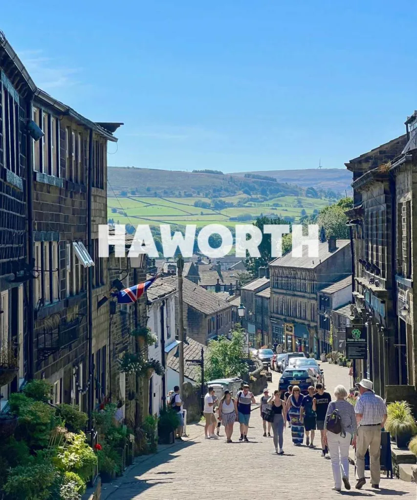 Steep cobblestone main street in Haworth village with stone buildings, tourists walking, and Yorkshire moors in background