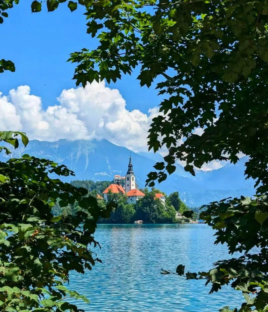 Picturesque view of Church of Assumption on Bled Island framed by tree branches with Julian Alps mountains in background, Slovenia