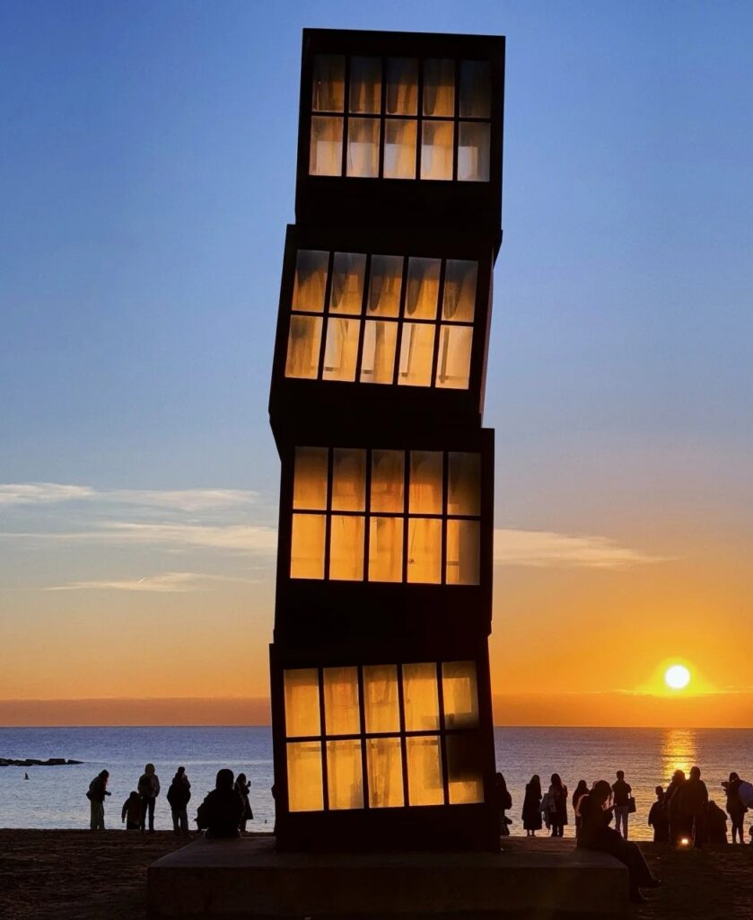 Modern beach sculpture silhouetted against orange sunset over Mediterranean Sea with crowds gathering at Barcelona's coastline