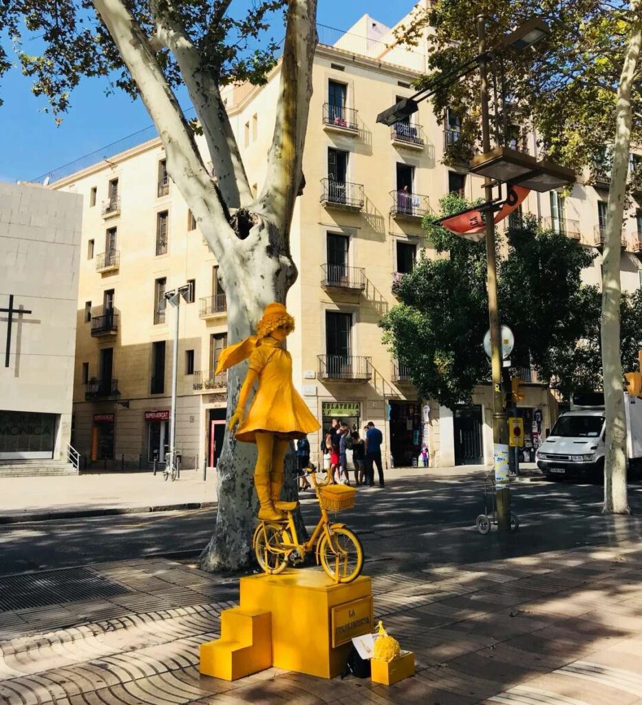 Street artist in bright yellow costume performing with bicycle in Barcelona plaza surrounded by traditional Catalan architecture