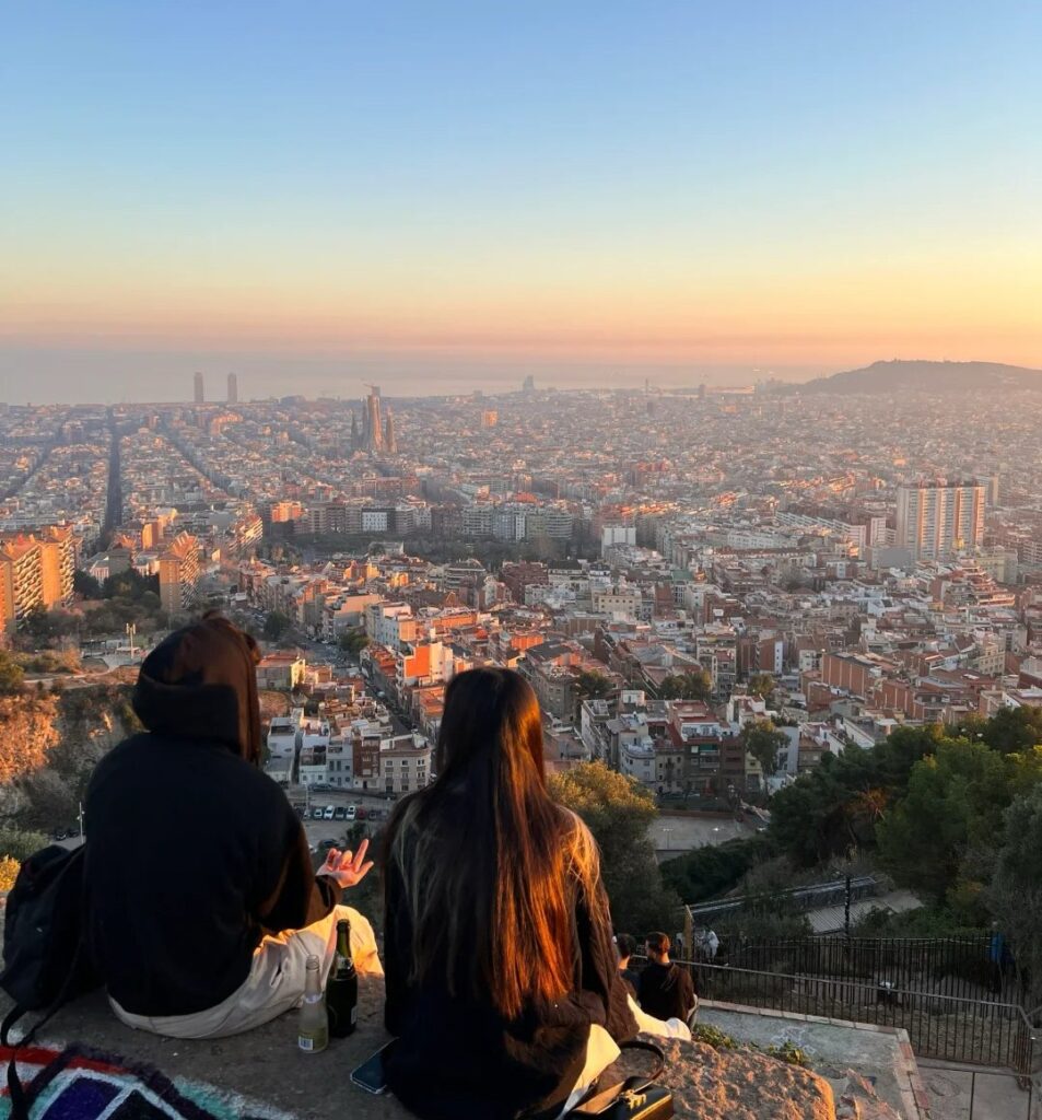 Two people watching golden hour sunset over Barcelona city skyline from elevated viewpoint with panoramic urban landscape