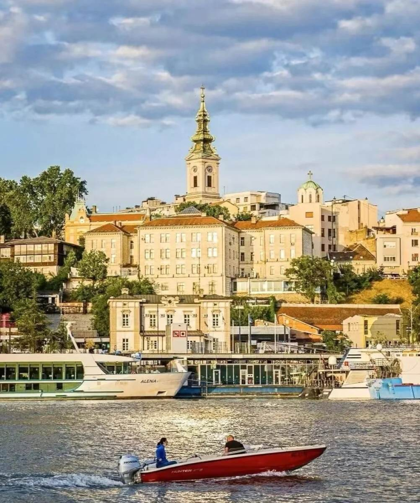 Belgrade cityscape showing Orthodox church spires and historic buildings along riverfront with tourist boats and small fishing vessel