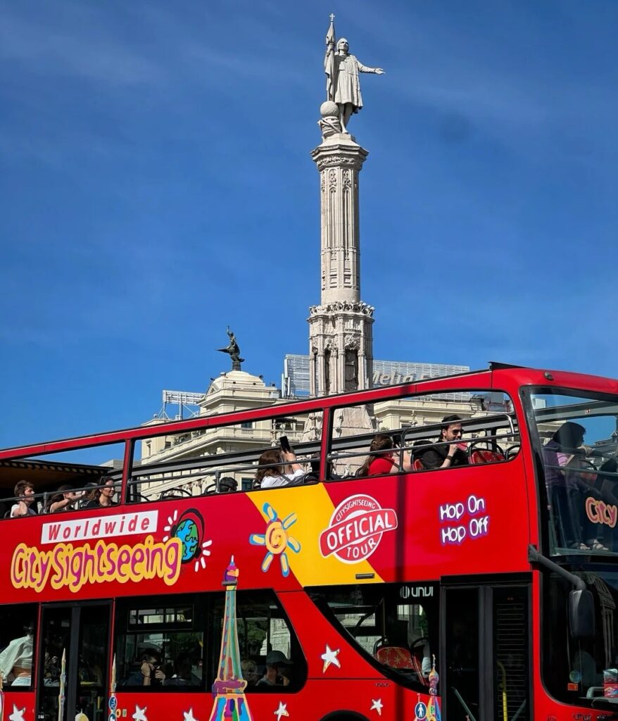 Red double-decker sightseeing bus at Madrid's Columbus Square with monument and tourists enjoying city tour