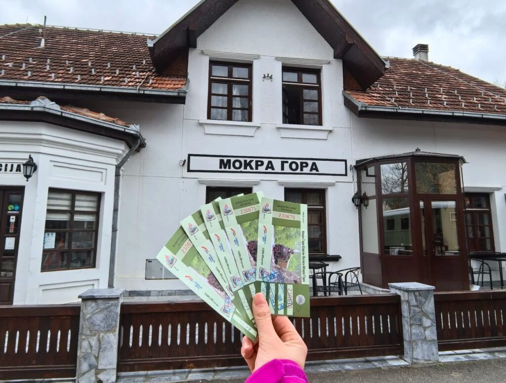 Hand holding multiple Šargan Eight railway tickets in front of historic Mokra Gora train station building with traditional architecture