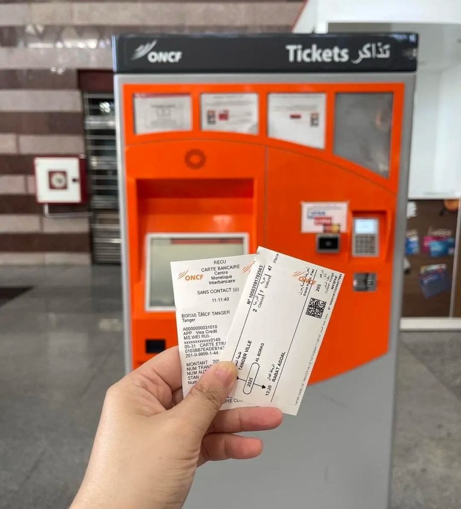 Hand holding ONCF train tickets in front of orange automated ticket machine at Moroccan railway station