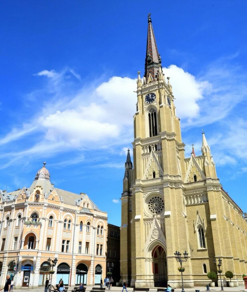 Neo-Gothic Catholic Cathedral of Name of Mary in Novi Sad with tall spire and ornate facade against blue sky with Austrian-Hungarian buildings nearby