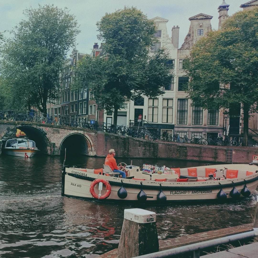 A tour boat cruising on an Amsterdam canal, with an old bridge, canal houses, and green trees in the background.
