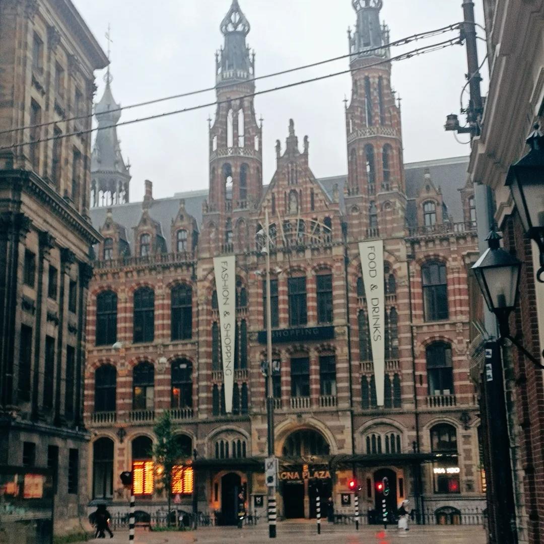 The exterior of the Magna Plaza building on a cloudy day. It is an ornate red-brick building with two flags hanging on the front.