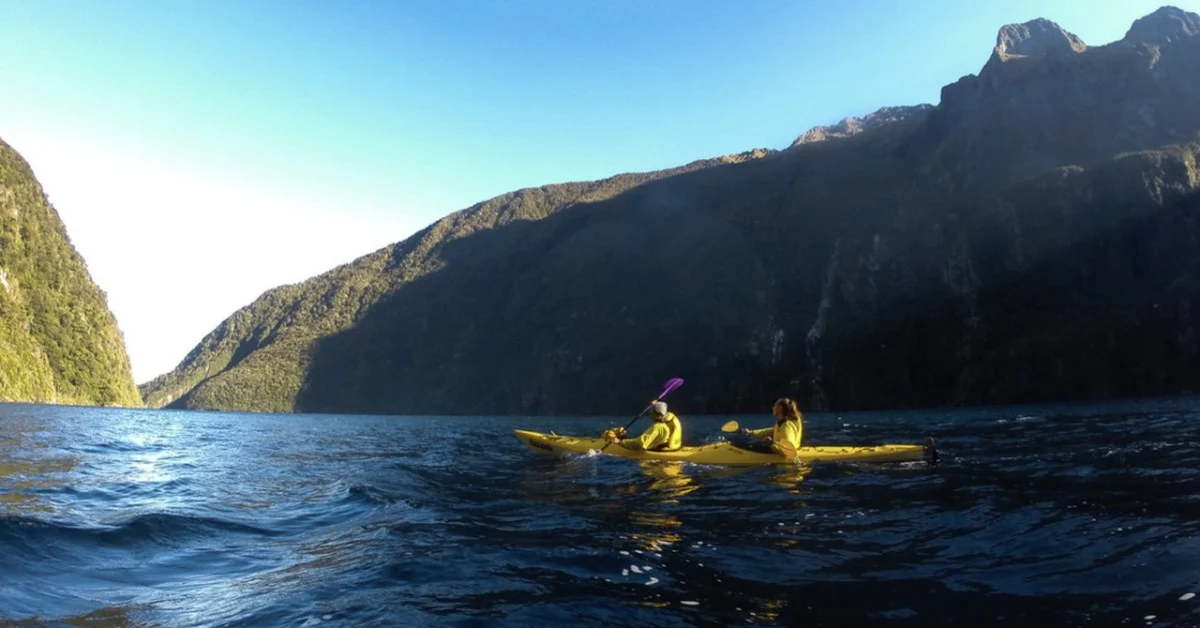 Panoramic aerial view of a large, blue, winding lake (Lake Wakatipu) surrounded by green and brown mountains under a partially cloudy sky in New Zealand.