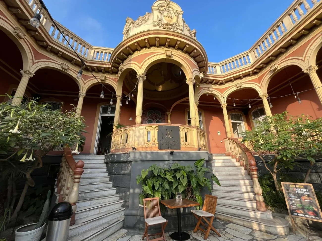 The grand, ornate entrance of Kokopelli Hostel Barranco in Lima, Peru, featuring a reddish building with a balcony, arches, and a grand staircase leading up to the main door. Two small tables with chairs are in the foreground.