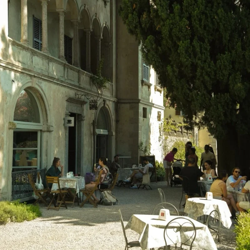 A group of people enjoying drinks and conversation at an outdoor cafe terrace with tables and chairs on a gravel ground, in front of an old, grand building with columns and arches, partially shaded by a large tree.