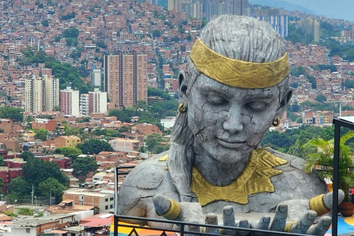 A massive weathered stone statue of an indigenous figure with a gold headband, overlooking the sprawling hills and houses of Medellín, Colombia.