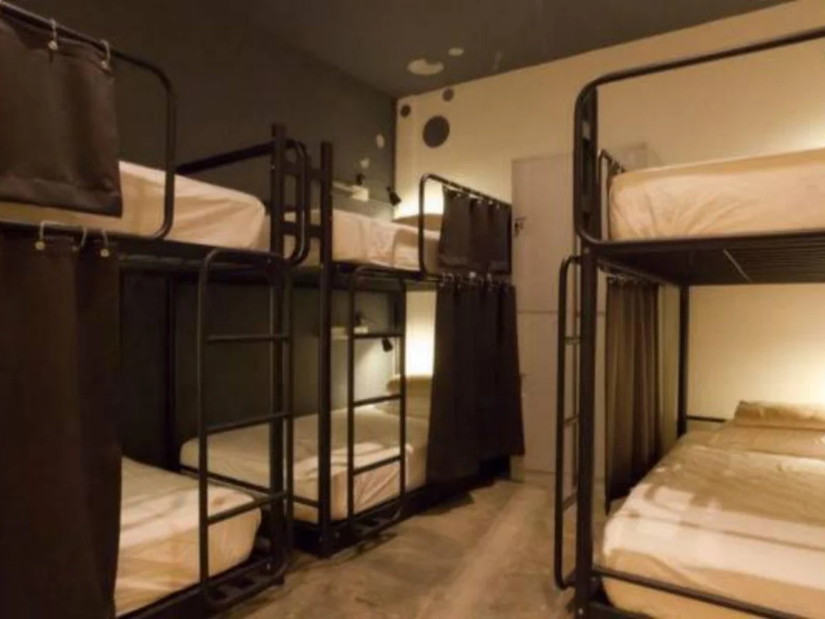 Clean modern hostel dormitory room featuring black metal bunk beds with white bedding, individual privacy curtains, reading lights, and personal storage lockers - typical of budget accommodations in Singapore