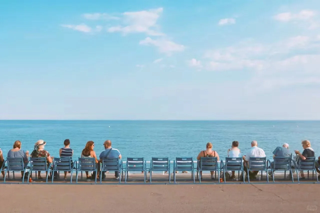 A line of people seated on blue chairs with their backs to the camera, facing the vast blue sea and clear sky on a sunny day in Nice, France.