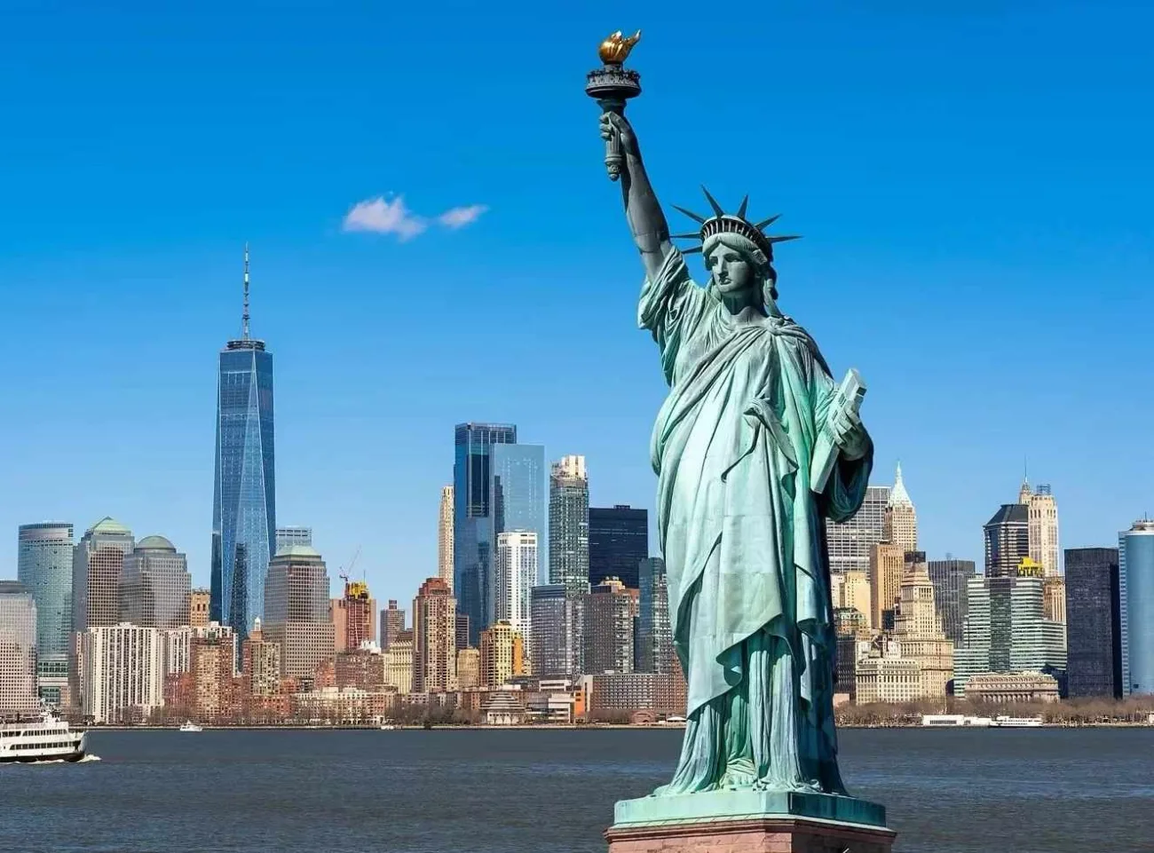 The Statue of Liberty standing prominently in the foreground, with the iconic Lower Manhattan skyline and One World Trade Center across the water under a clear blue sky.