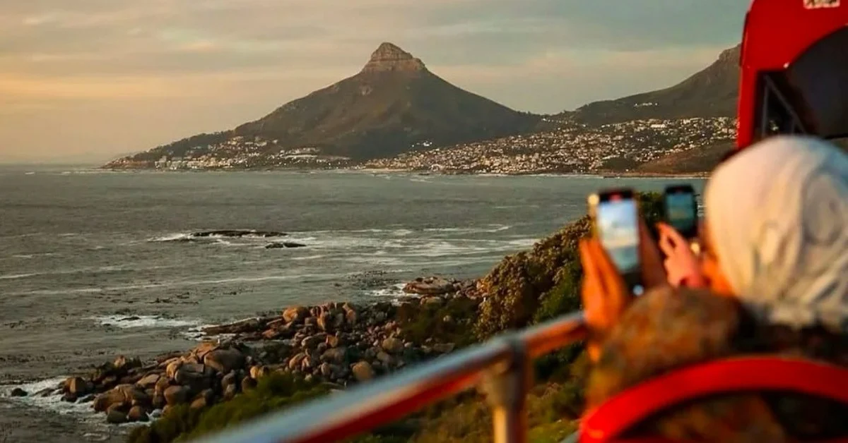 A person on a tour bus photographing Lion's Head mountain and the coastline of Cape Town, South Africa, at sunset.